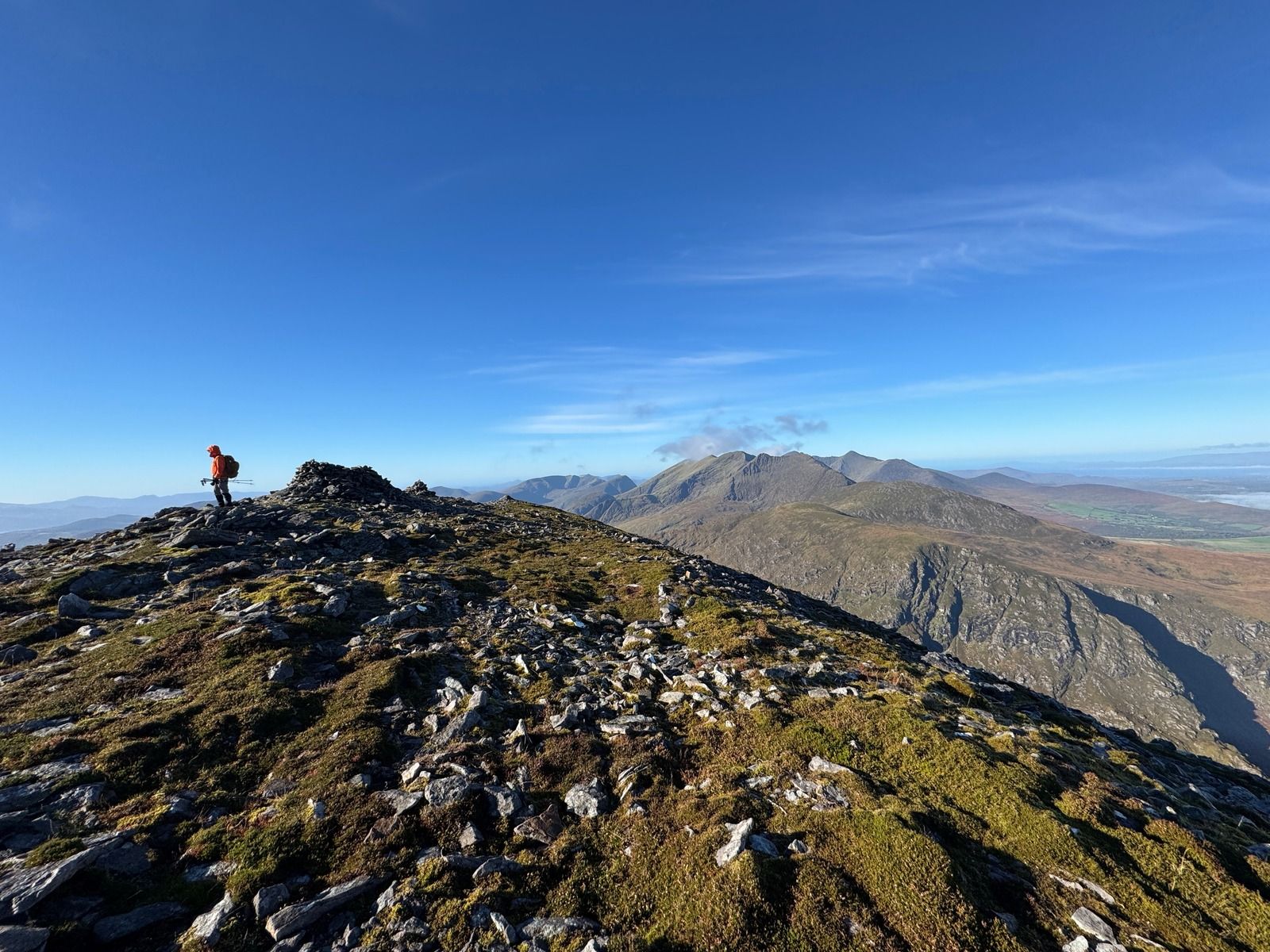 A walker standing beside a cairn on a slate summit, with mountains beyond