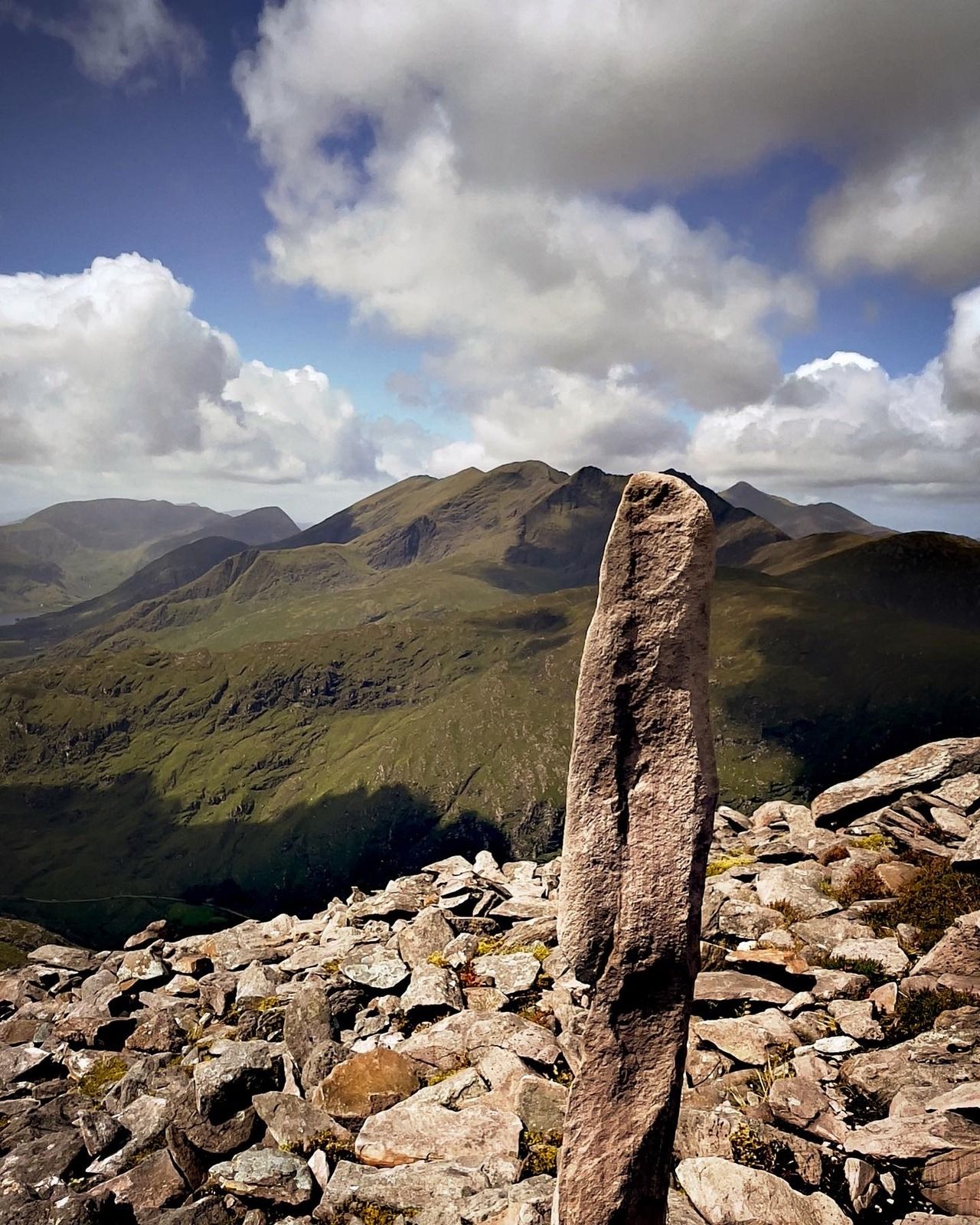 A tall upright stone slab set in a cairn, with green mountains and cloud beyond