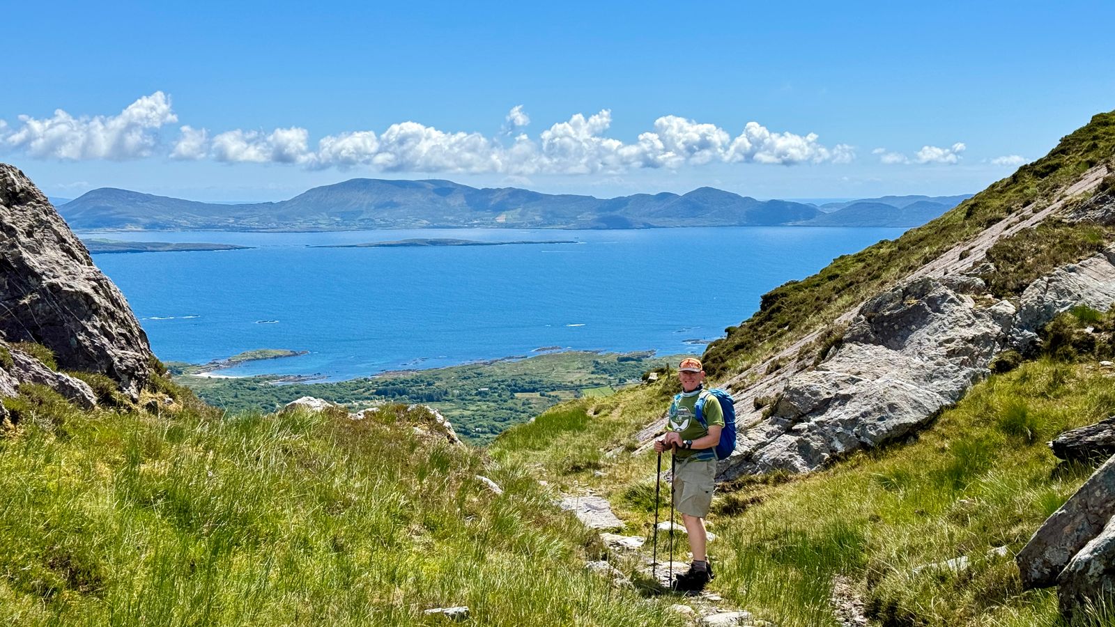A walker with poles on a grassy path between rocks, with a broad blue bay and distant hills beyond