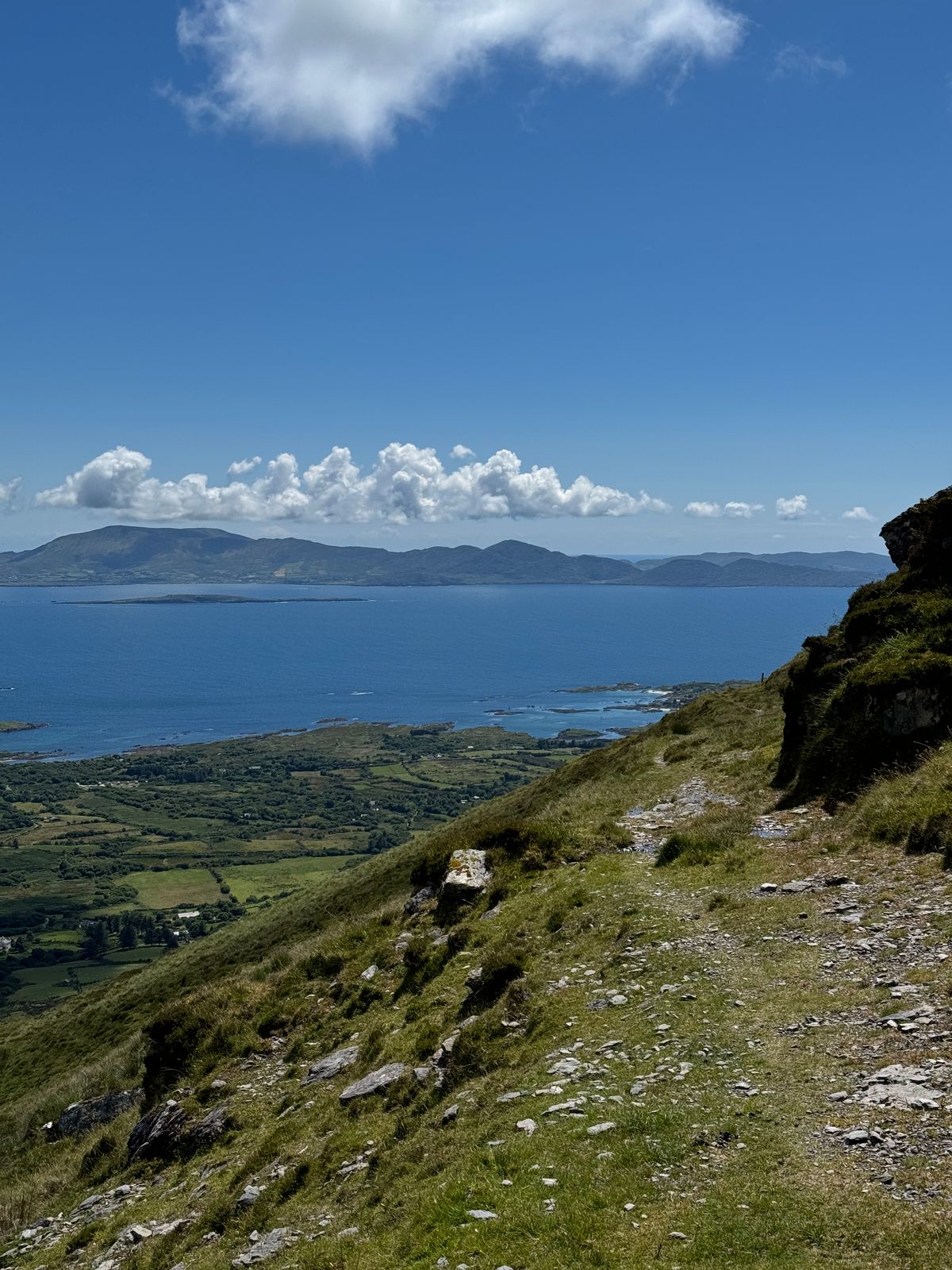 A gravel and rock trail on the hillside above blue water and distant mountains