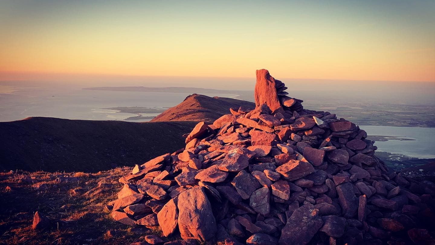 A stone summit cairn lit orange by the setting sun above the coast