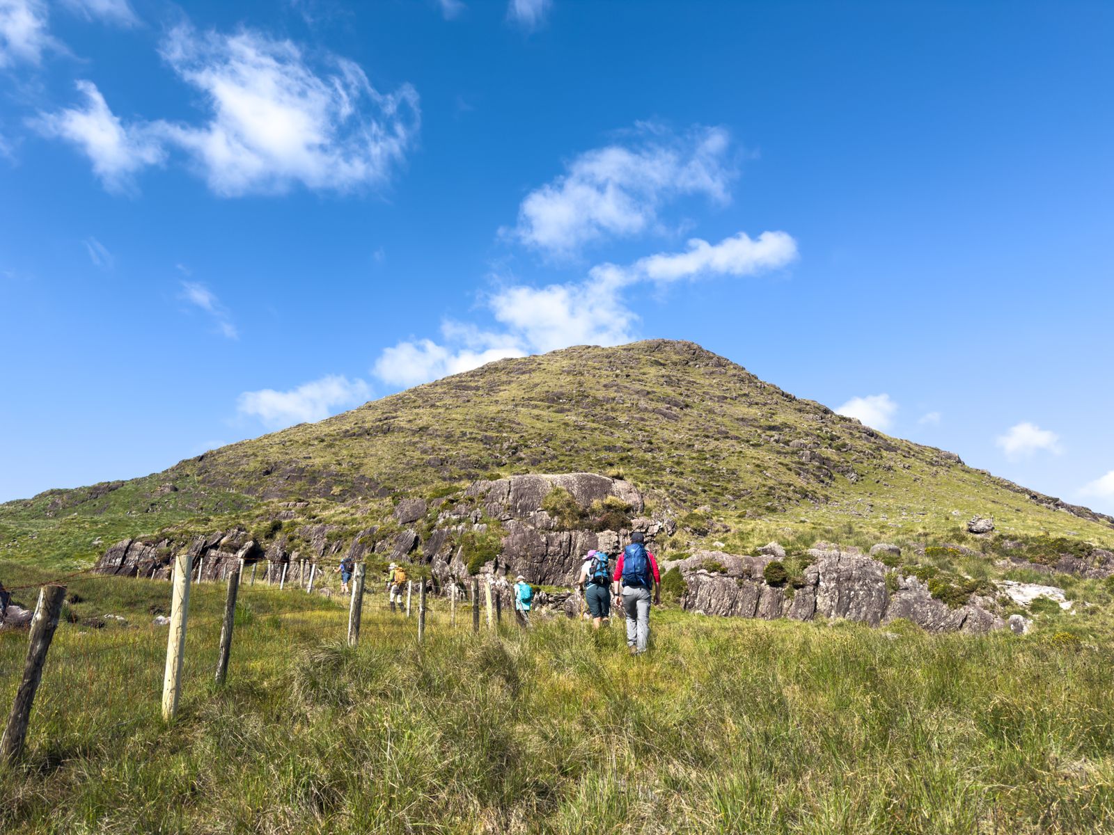 Walkers approaching a rounded grassy top of Cnoc na gCapall beneath a vivid blue sky