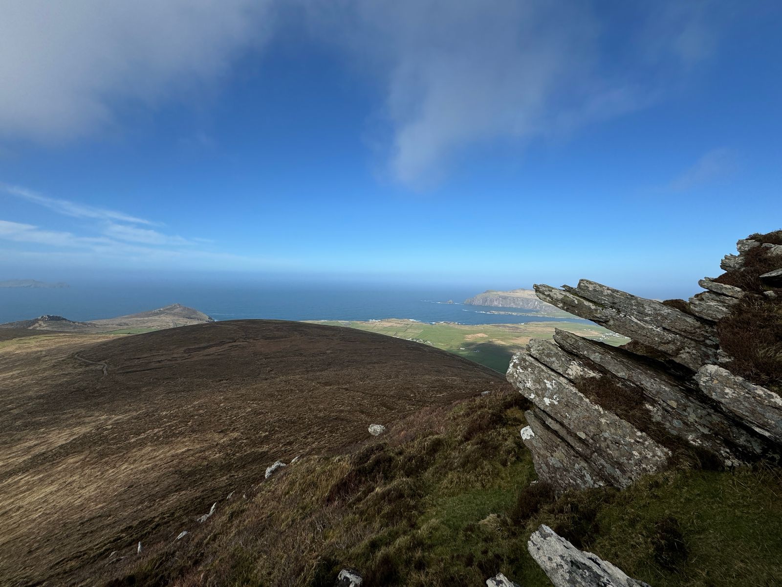 Tilted rock slabs above a rounded grassy ridge, coastal fields and the blue Atlantic