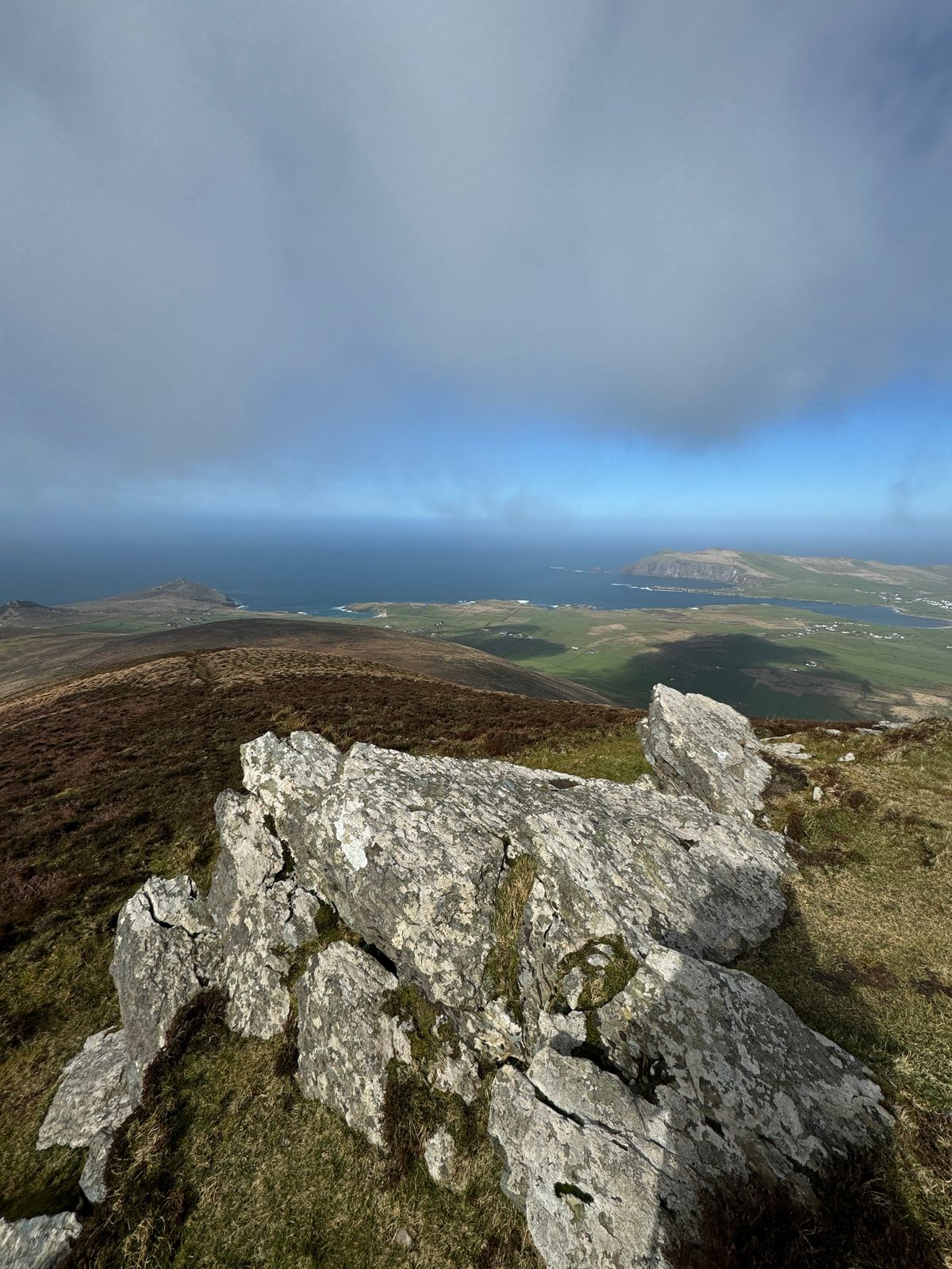 Lichen-covered rocks overlooking fields and coastline beneath low cloud