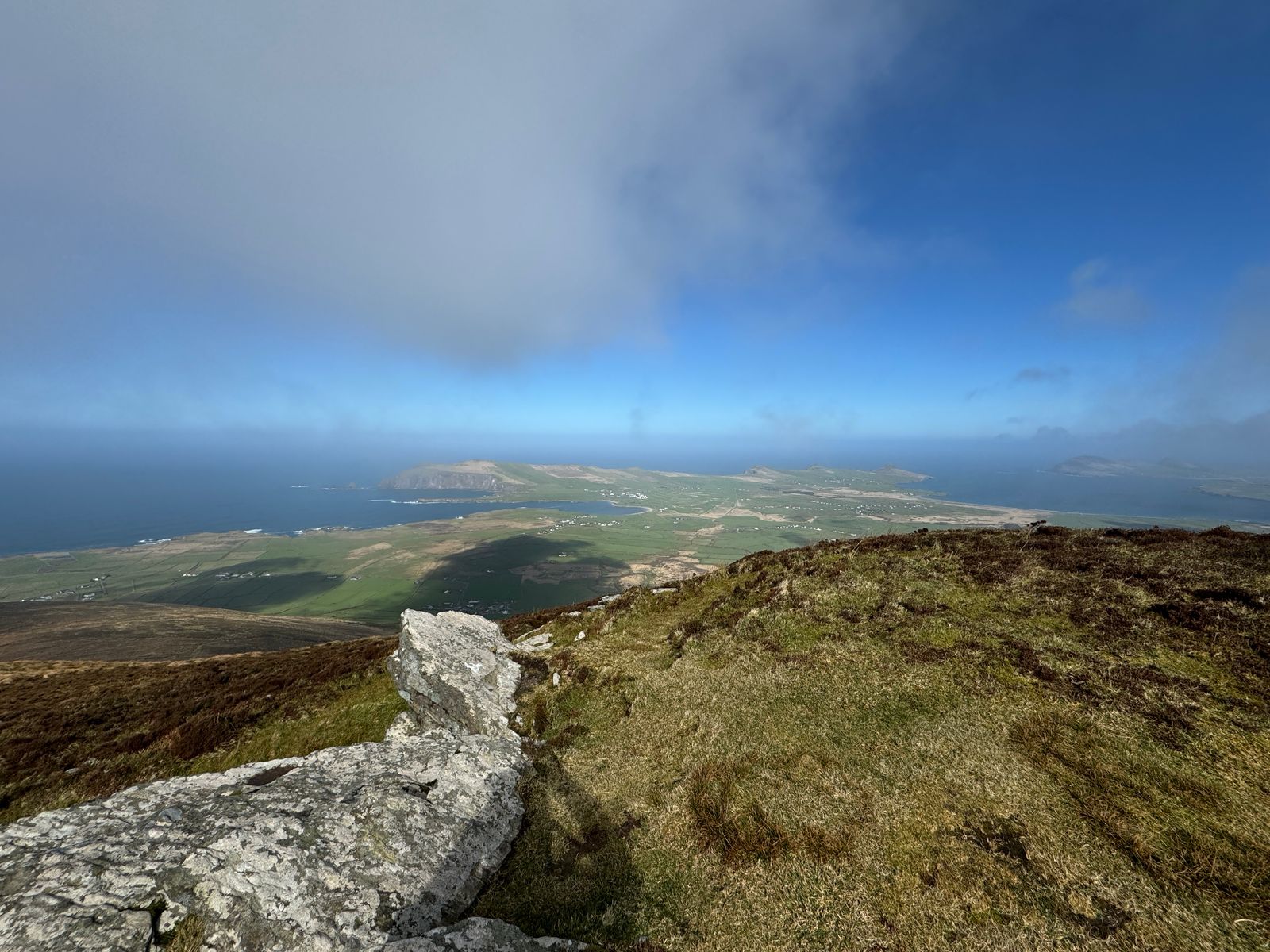 Rocky hillside above patchwork fields and a long coastline under clearing cloud
