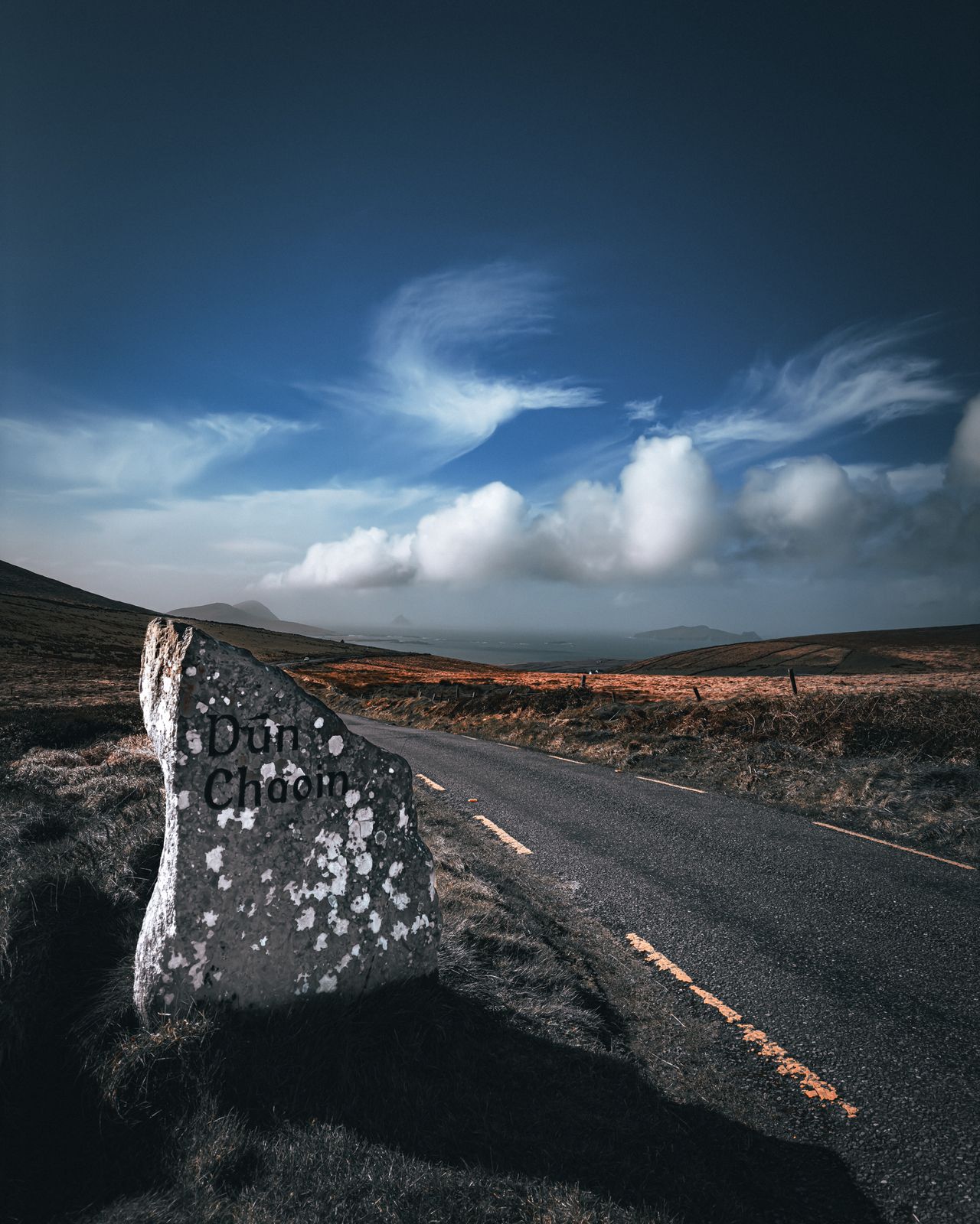 A roadside stone marked Dún Chaoin beside a road descending towards the Atlantic