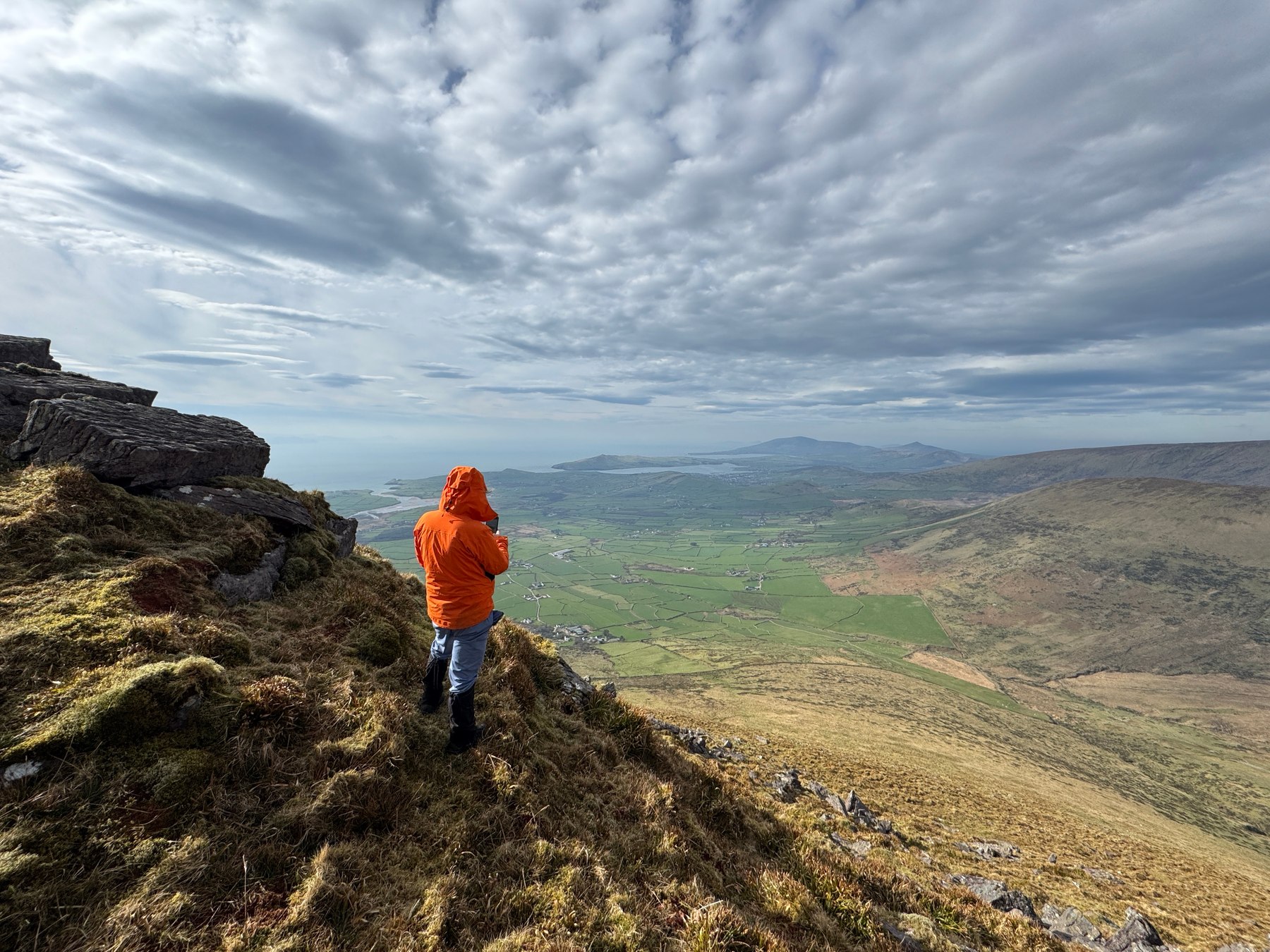 A walker descending rough ground towards green fields far below