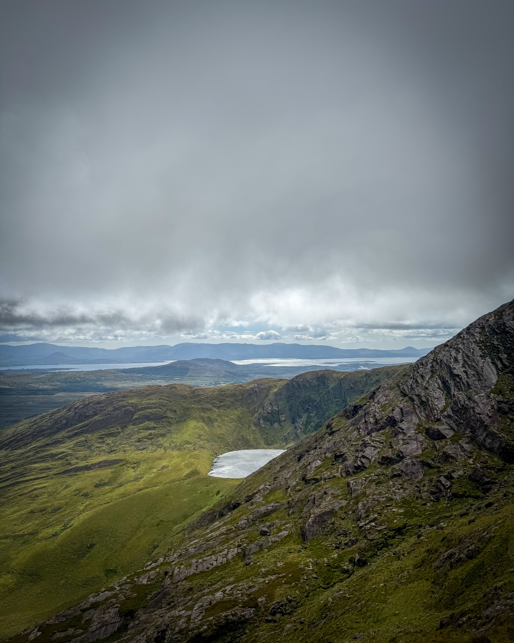 A small mountain lough below a dark ridge with coastal water in the distance