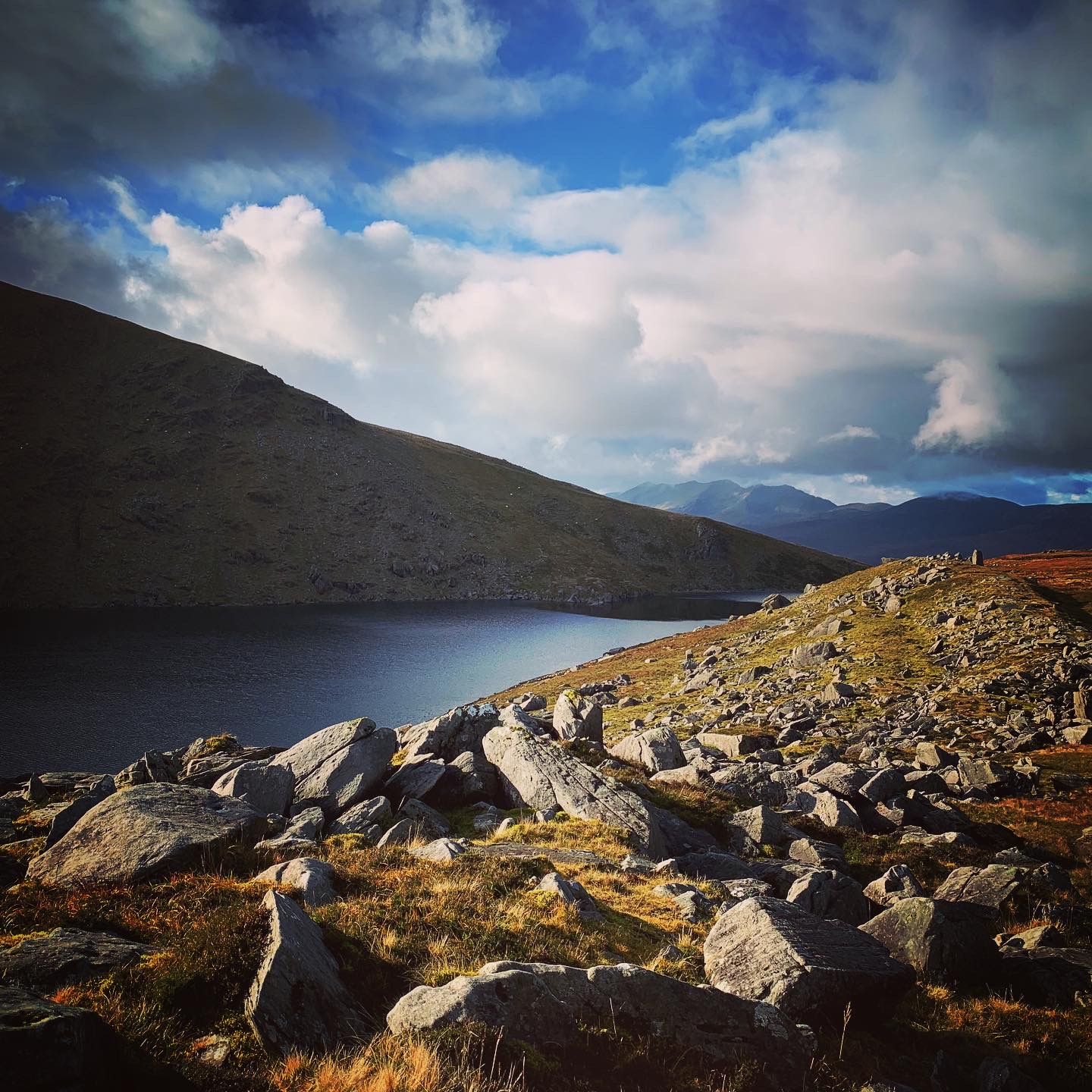 Angular rocks and golden grass beside a mountain lake under a blue and cloudy sky