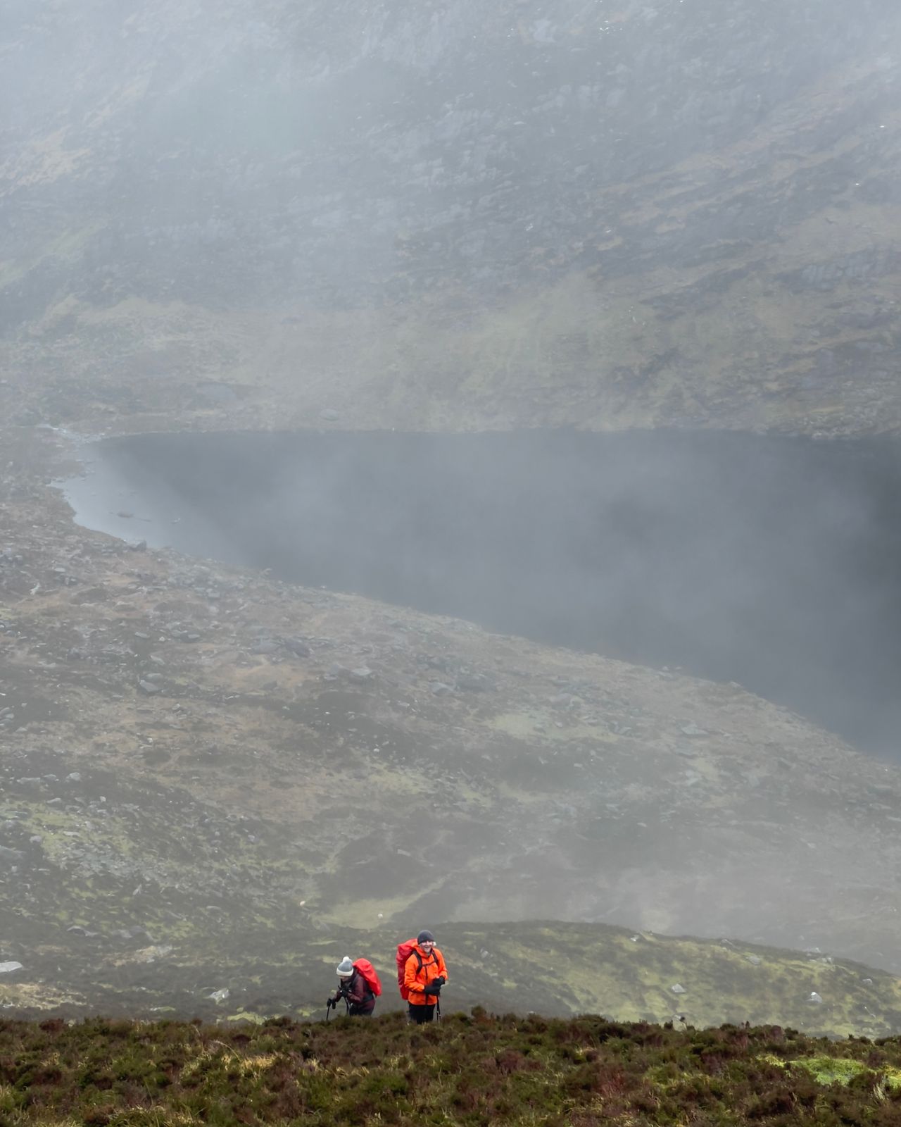 Two walkers in red jackets below a lake and rocky cliffs partly hidden by mist