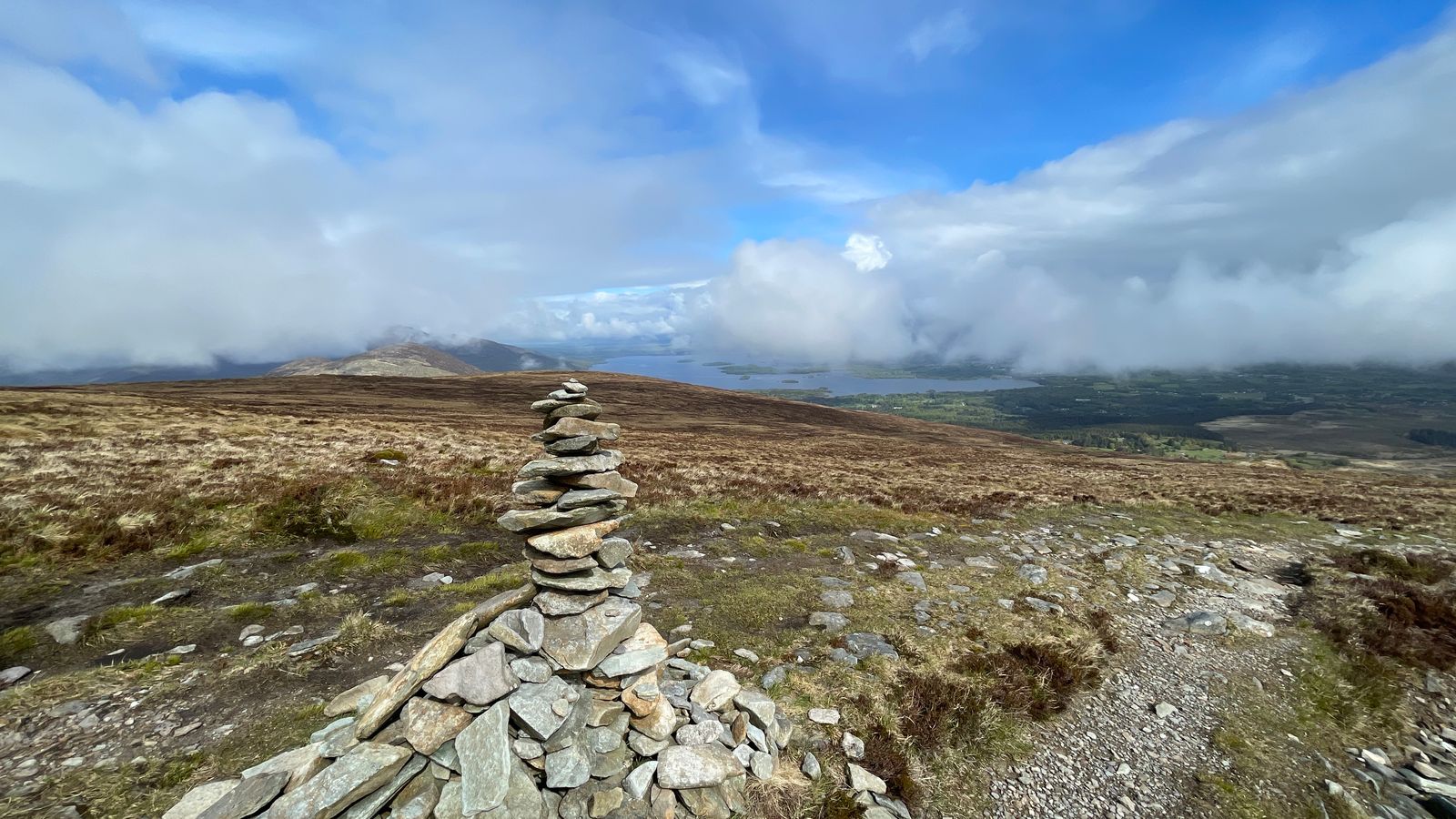 A narrow stone cairn on open moorland beneath blue sky and low cloud