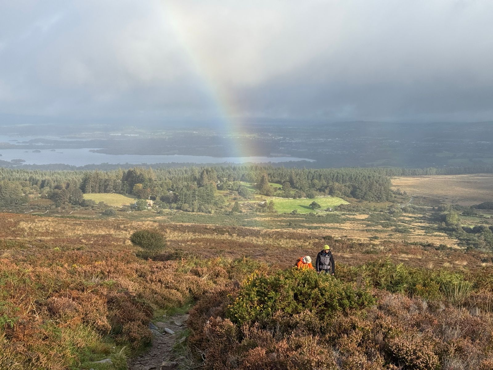 Two walkers on a heather-covered hillside below a faint rainbow