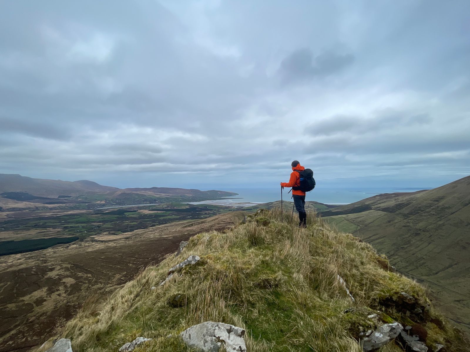 A walker on a grassy mountain shoulder looking north towards the coast and Tralee Bay