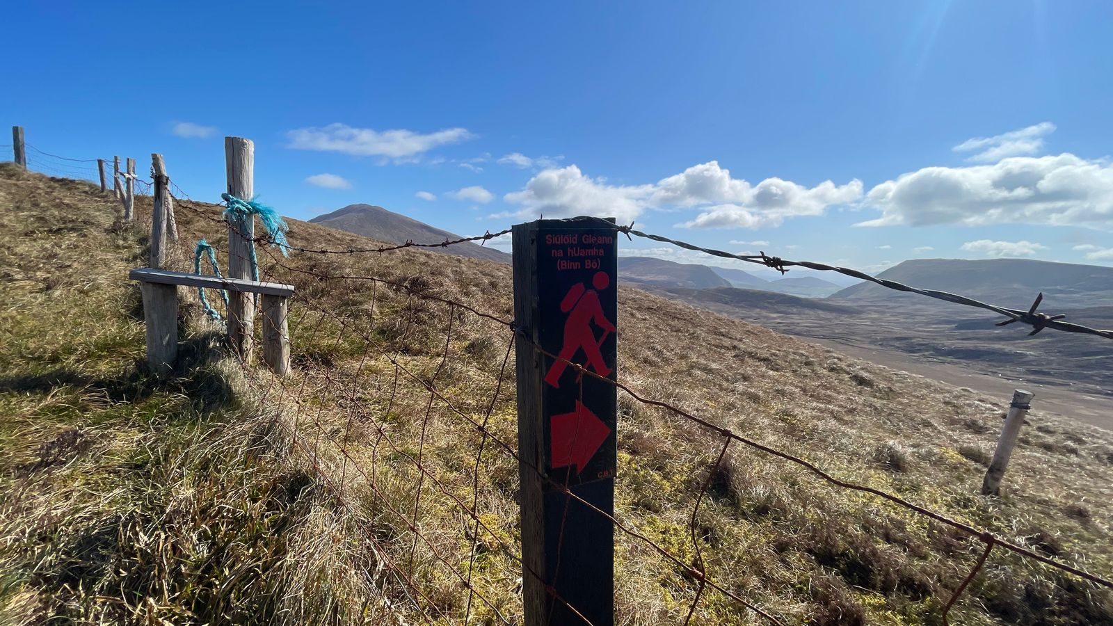 A Glenahoo walking-trail waymarker beside a fence with mountains in the distance