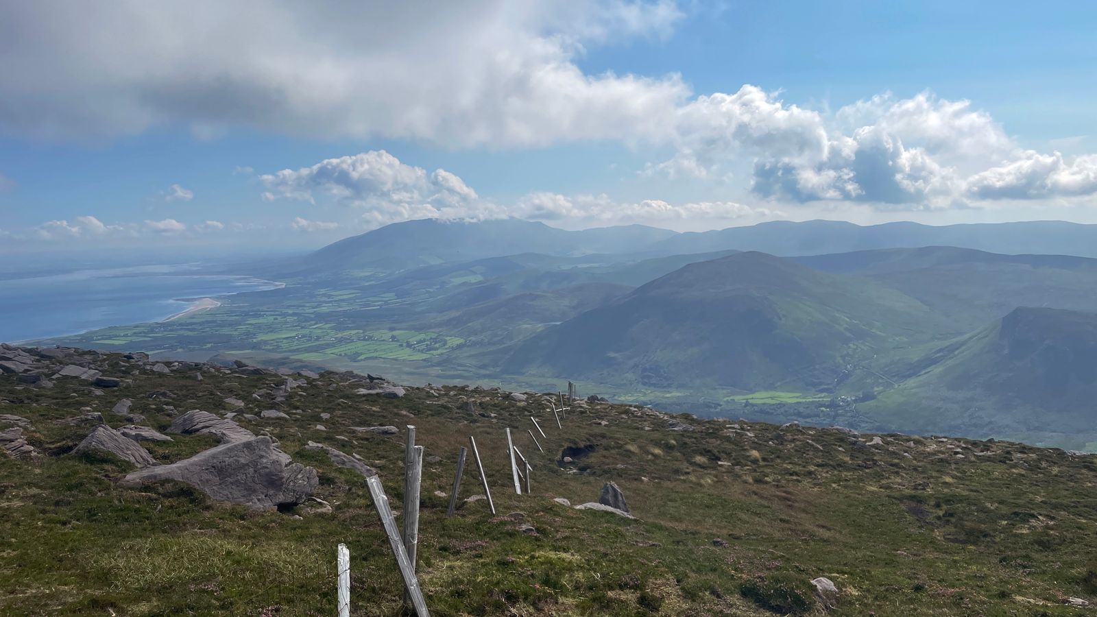 A fence running down rough mountain ground towards a wide view of the hills and coast